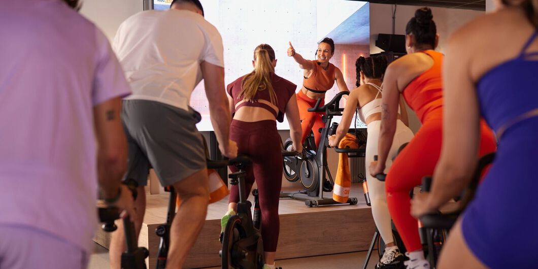 People ride stationary bikes in a group while an instructor on a raised platform gives a thumbs-up gesture during the class.