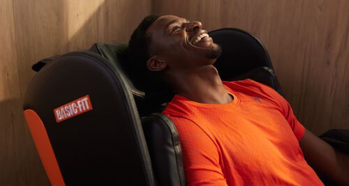 A man in an orange sports shirt relaxes and smiles while sitting in a black Basic-Fit massage chair. 
