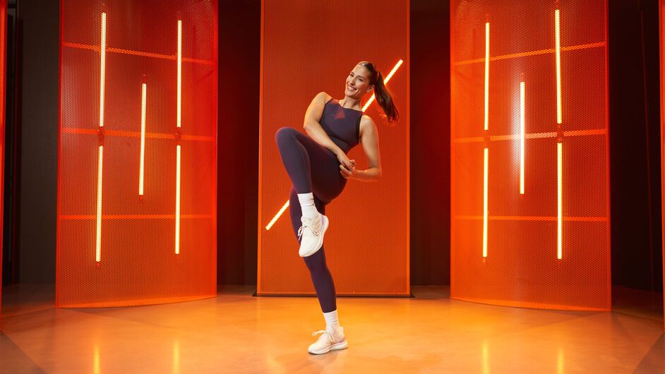 A woman balances on one leg while lifting the other knee, performing a controlled movement in a fitness studio with orange lighting panels in the background.