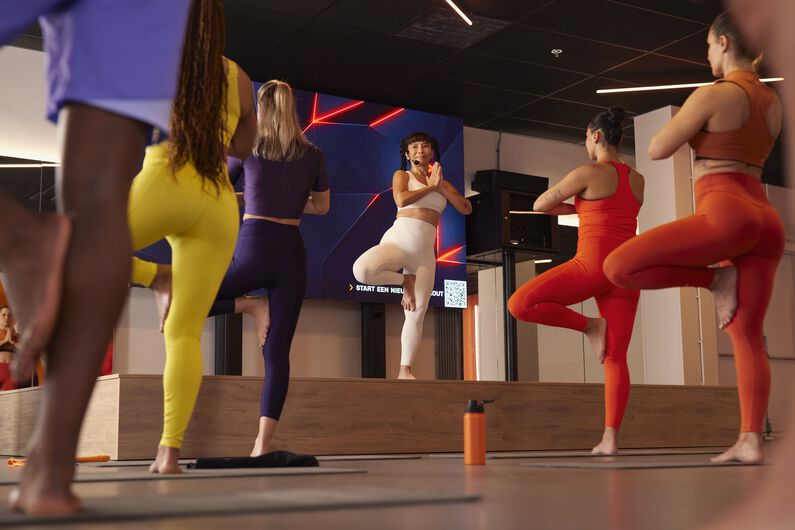 Yogatrainerin leitet eine Gruppenstunde von einer B&uuml;hne aus, w&auml;hrend die Teilnehmenden auf einem Bein balancieren