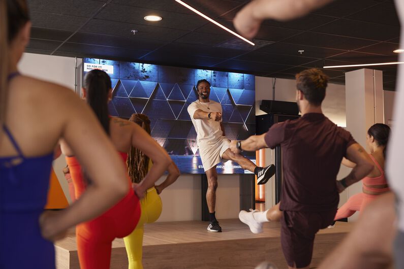 Group fitness class in a Basic-Fit studio, with an instructor on a stage demonstrating a movement while participants exercise in front of a screen.