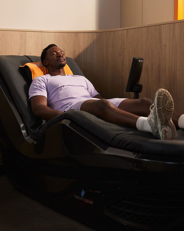 A man in a light purple sports outfit relaxes on a hydromassage bed with an orange towel behind his head at Basic-Fit.