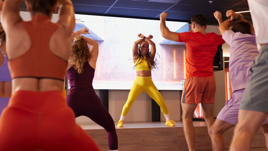 A group follows a high-energy workout, raising their arms and moving in sync while an instructor leads from a stage in a fitness studio.