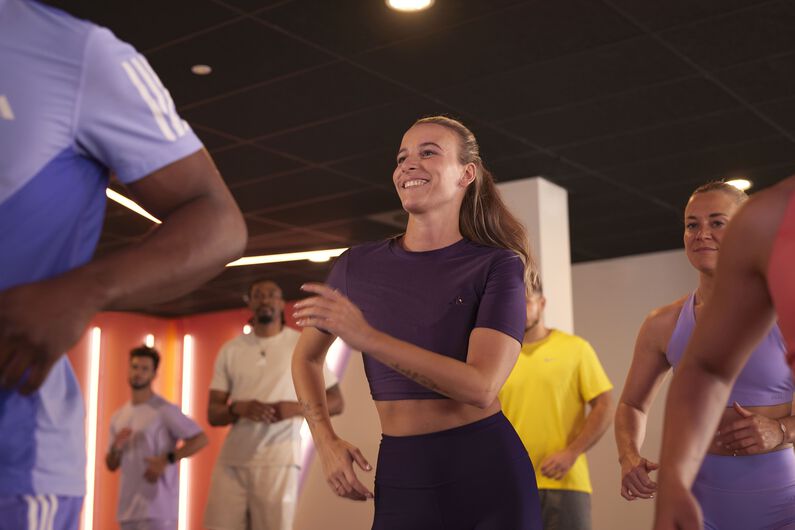A group of people performs coordinated dance movements while facing the front of the studio during a group workout.