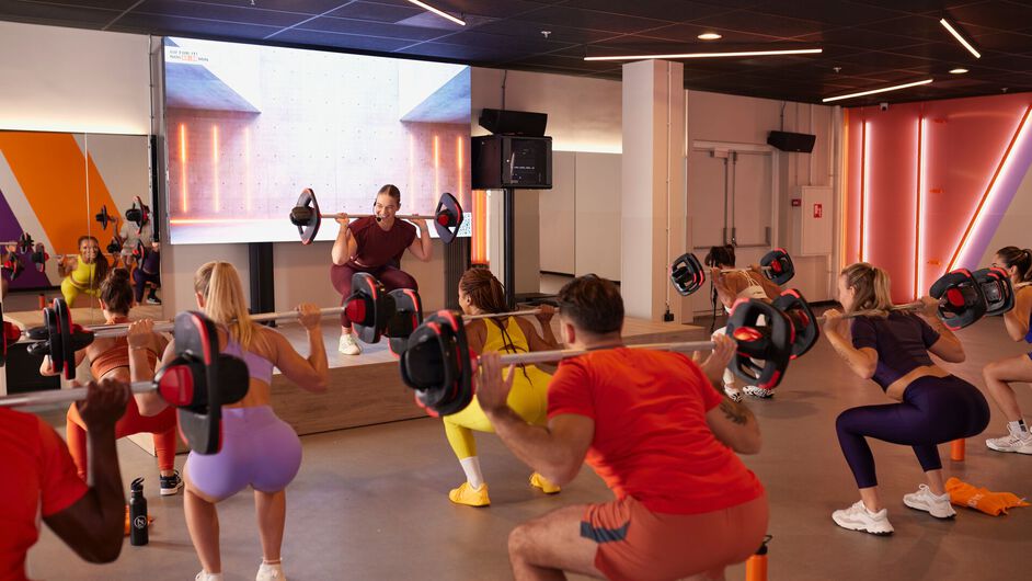 a group of people performs squats with barbells while an instructor demonstrates the movement on a raised stage in a fitness studio