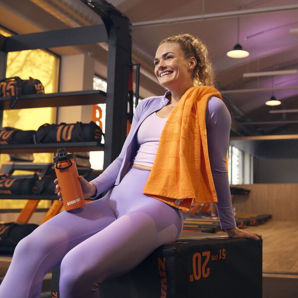 Frau in violetter Sportbekleidung, die auf einer plyometrischen Box in einem Basic-Fit Fitnessstudio sitzt, eine orangefarbene Basic-Fit Trinkflasche h&auml;lt und ein oranges Handtuch &uuml;ber der Schulter tr&auml;gt.