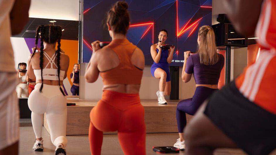 Participants hold weight plates while performing lunges as an instructor demonstrates the exercise on a stage in a fitness studio.