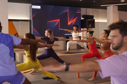 Gruppe von Personen, die an einem yoga&auml;hnlichen Gruppenkurs in einem Fitnessstudio teilnehmen, in einer breiten stehenden Haltung mit ausgestreckten Armen, angeleitet von einer Trainerin auf einer Plattform.
