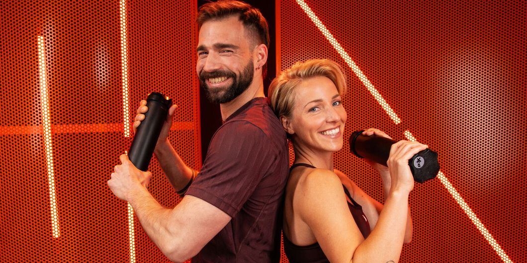 Twee personen die rug aan rug in een fitnessstudio staan, lachend met sportwaterflessen in hun handen.