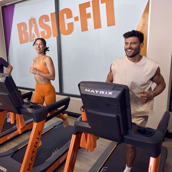 Zwei Personen laufen nebeneinander auf Laufb&auml;ndern in einem Basic-Fit Studio und l&auml;cheln dabei. Das orangefarbene Basic-Fit Logo ist an der Wand hinter ihnen zu sehen.