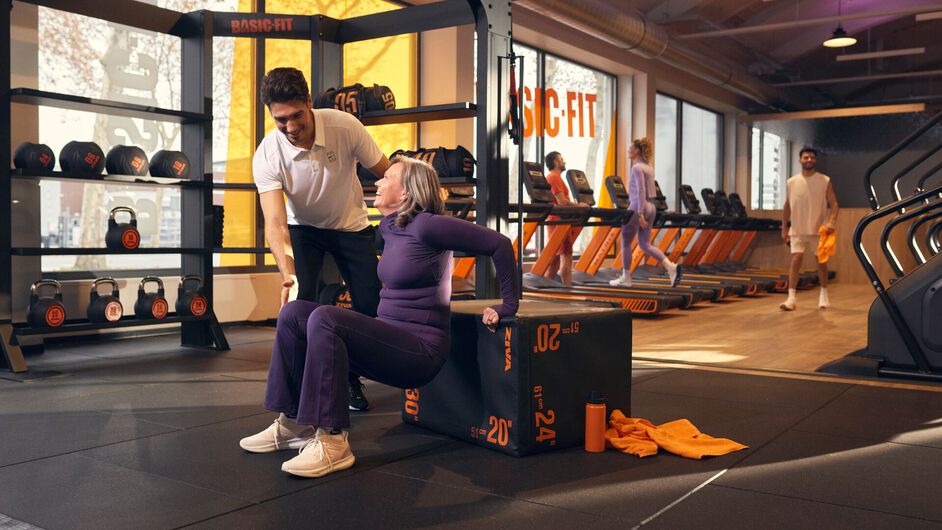 Un fisioterapeuta acompa&ntilde;a a una mujer mayor durante un ejercicio de fuerza sentado sobre una plyobox en un gimnasio Basic-Fit. Otros miembros entrenan en m&aacute;quinas de cardio al fondo.