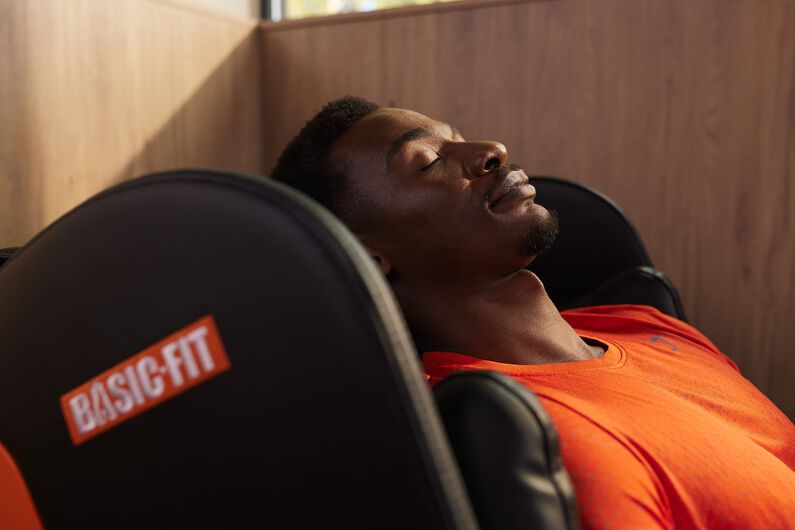 A man in an orange sports shirt relaxes with his eyes closed in a black massage chair with the Basic-Fit logo.