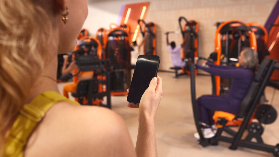 Person holding a smartphone in a Basic-Fit gym, using the app to book a group class while other members train on strength machines in the background.