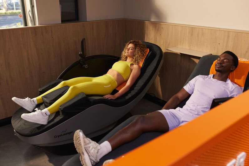 A woman in a yellow sports outfit relaxes on a cryo bed, while a man in a light purple outfit rests on a hydromassage bed at Basic-Fit.
