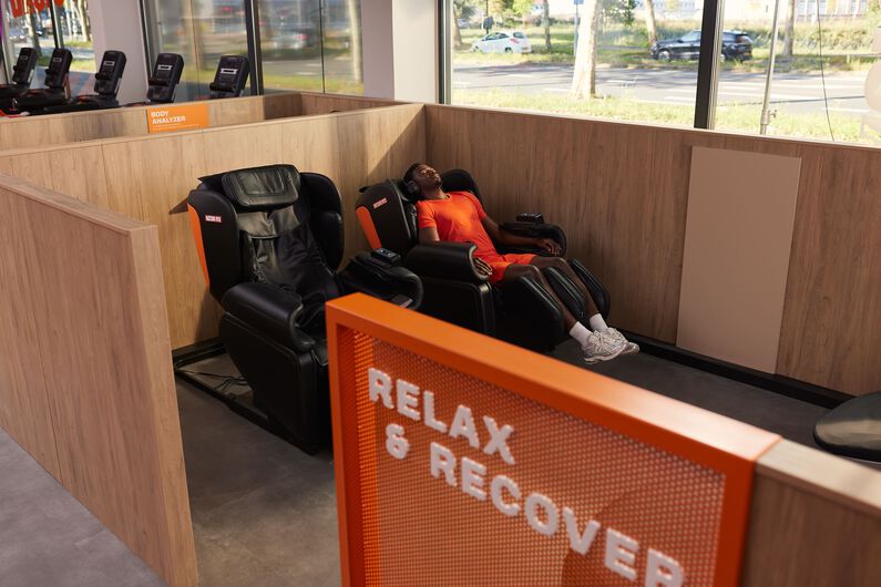 A man in a red sports outfit relaxes with headphones in a massage chair in the Relax & Recover zone at Basic-Fit.