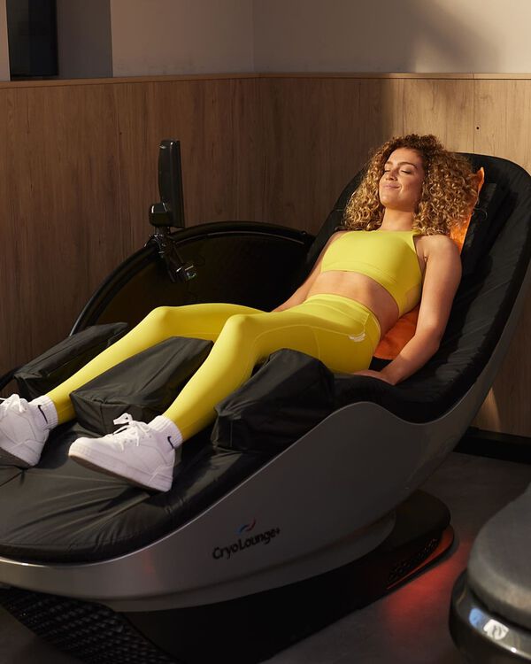 A woman in a yellow sports outfit relaxes on a CryoLounge+ recovery chair with an orange towel behind her head at Basic-Fit.