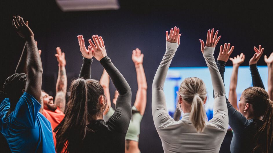 Un grupo de miembros sigue juntos un entrenamiento en v&iacute;deo en Basic-Fit y levanta los brazos de forma sincronizada mientras mira la pantalla en el estudio.