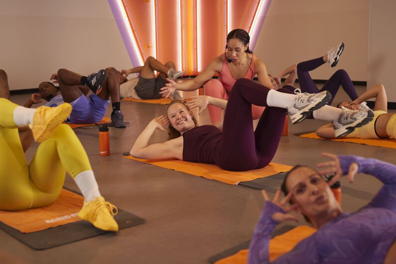 Abs & Core groepsles op matten, met een instructeur die een deelnemer begeleidt tijdens een core-oefening op de vloer.
