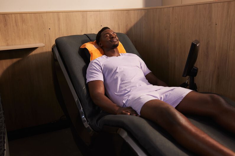 A man in a light purple sports outfit relaxes on a hydromassage bed with an orange towel behind his head at Basic-Fit.