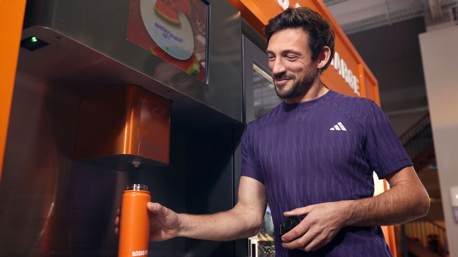 Mann in einem Fitnessstudio, der eine orangefarbene Basic-Fit Trinkflasche an einer Yanga Sports Water Station auff&uuml;llt und dabei l&auml;chelt.