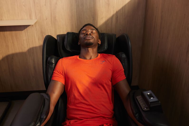 A man in a red sports outfit relaxes with his eyes closed in a massage chair at Basic-Fit.