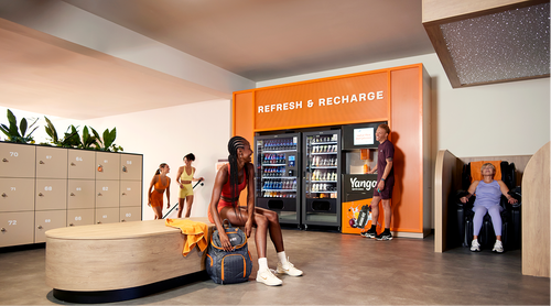 People relaxing and socializing in a modern gym lounge with lockers, a refreshment vending area, and seating, while members in workout clothing take a break before or after training.