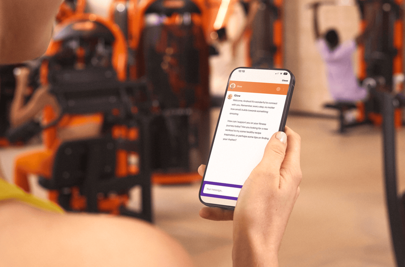 Person holding a smartphone in a Basic-Fit gym, using the app to get workout guidance while machines are visible in the background