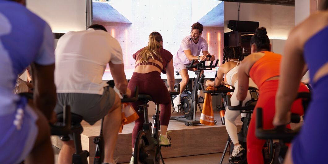 A group cycles on stationary bikes while an instructor on a raised platform leans forward on the handlebars and looks toward the participants.