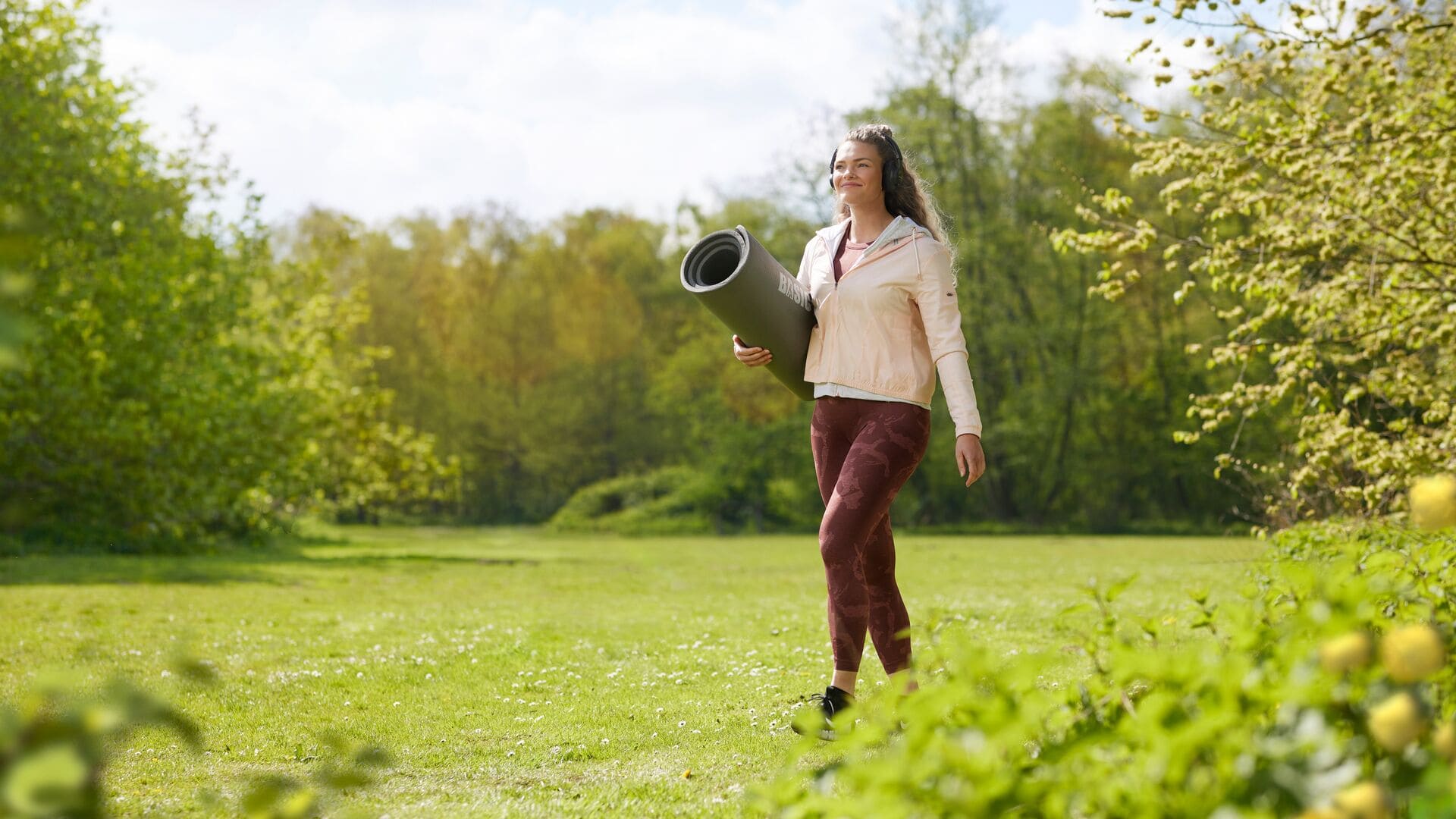 Eine Frau geht durch einen grünen Park mit einer zusammengerollten Yogamatte unter dem Arm, lächelt und trägt Sportkleidung.