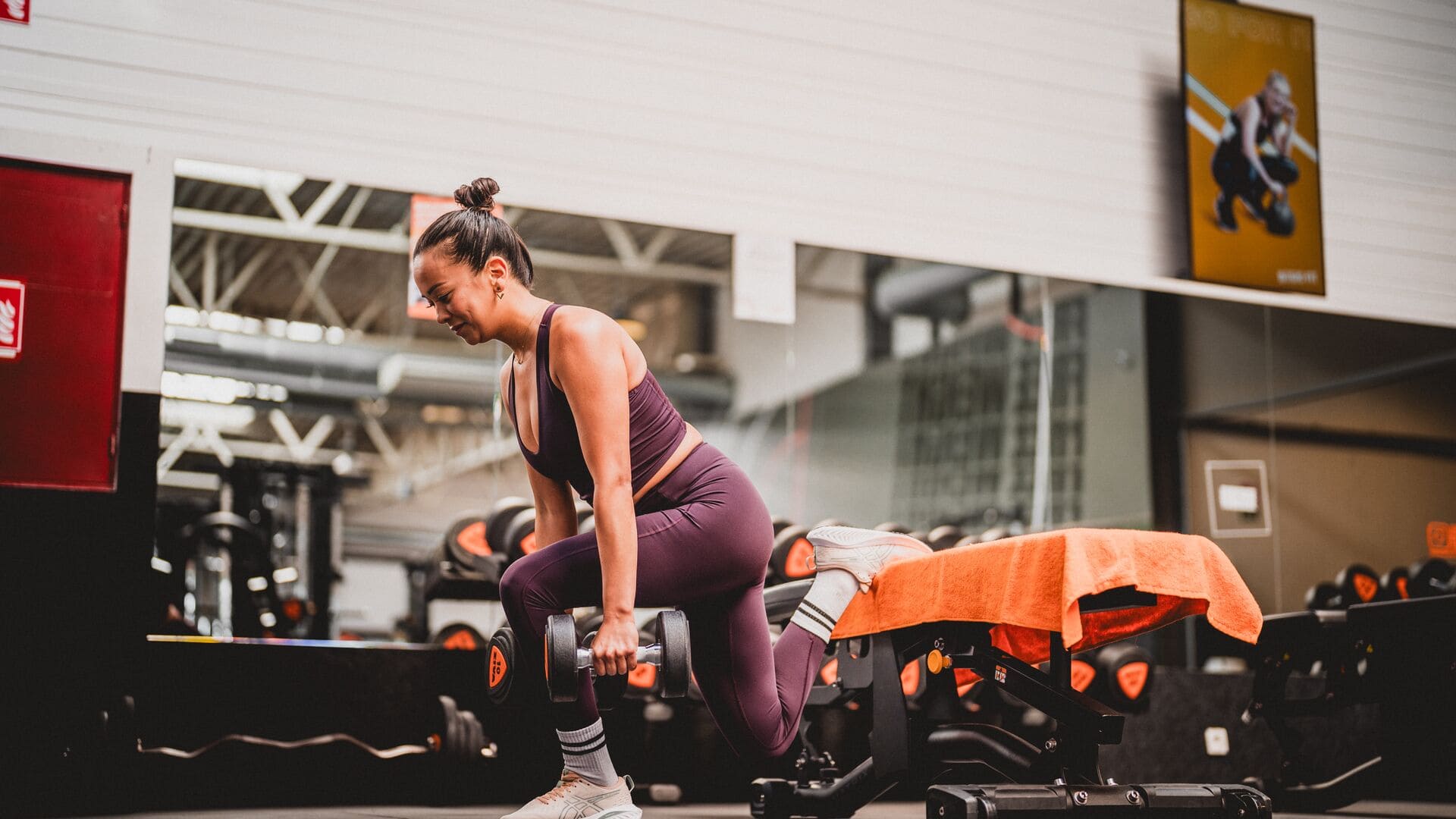 Frau in lilafarbener Sportkleidung führt einen Bulgarian Split Squat mit Kurzhanteln auf einer Bank im Basic-Fit Fitnessstudio aus.