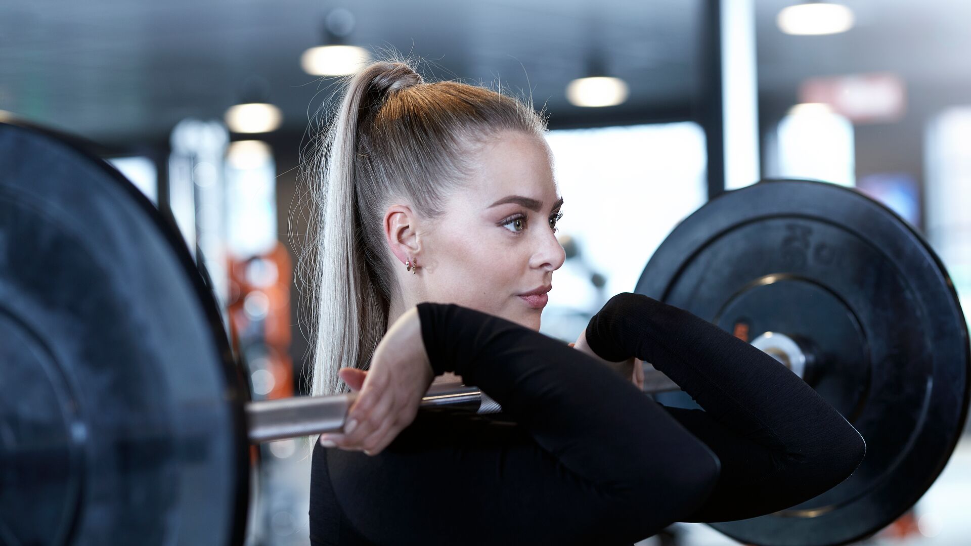 Woman holding a barbell in front rack position at a Basic-Fit gym. She is wearing a black long-sleeve top and focused on her lift, with strength equipment and soft lighting in the background.