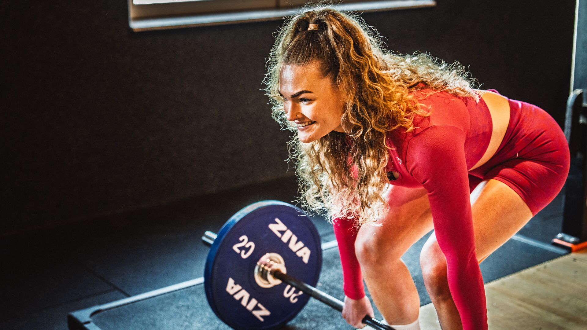 Eine Frau in roter Sportkleidung hebt in einem Basic-Fit-Fitnessstudio eine Langhantel und lächelt. Warmes Licht fällt von der Seite auf ihr Gesicht.