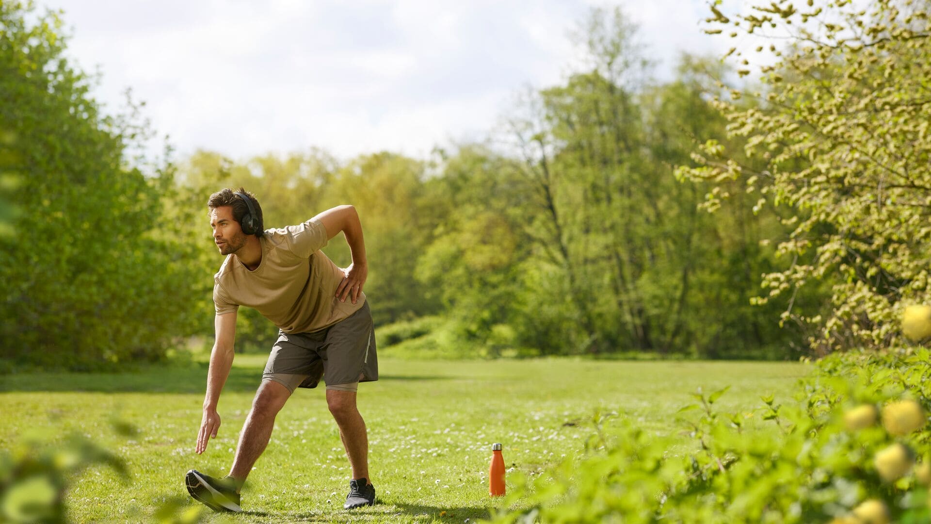 Ein Mann in einem beigen T-Shirt und Shorts dehnt sich draußen auf dem Gras, trägt Kopfhörer, mit einer orangefarbenen Trinkflasche neben sich.