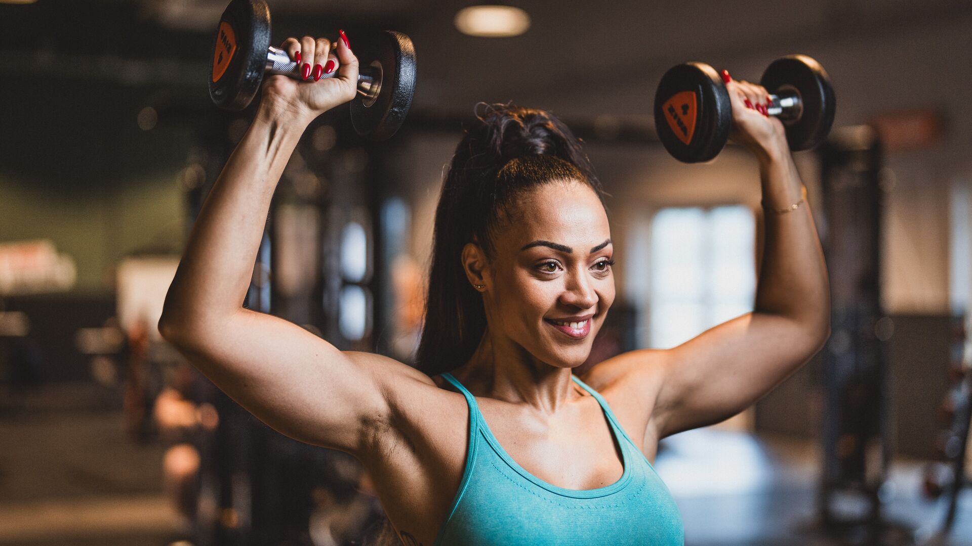 Woman lifting dumbbells overhead in a Basic-Fit gym, wearing a turquoise workout top. She is smiling and focused, with gym equipment and natural light visible in the background.