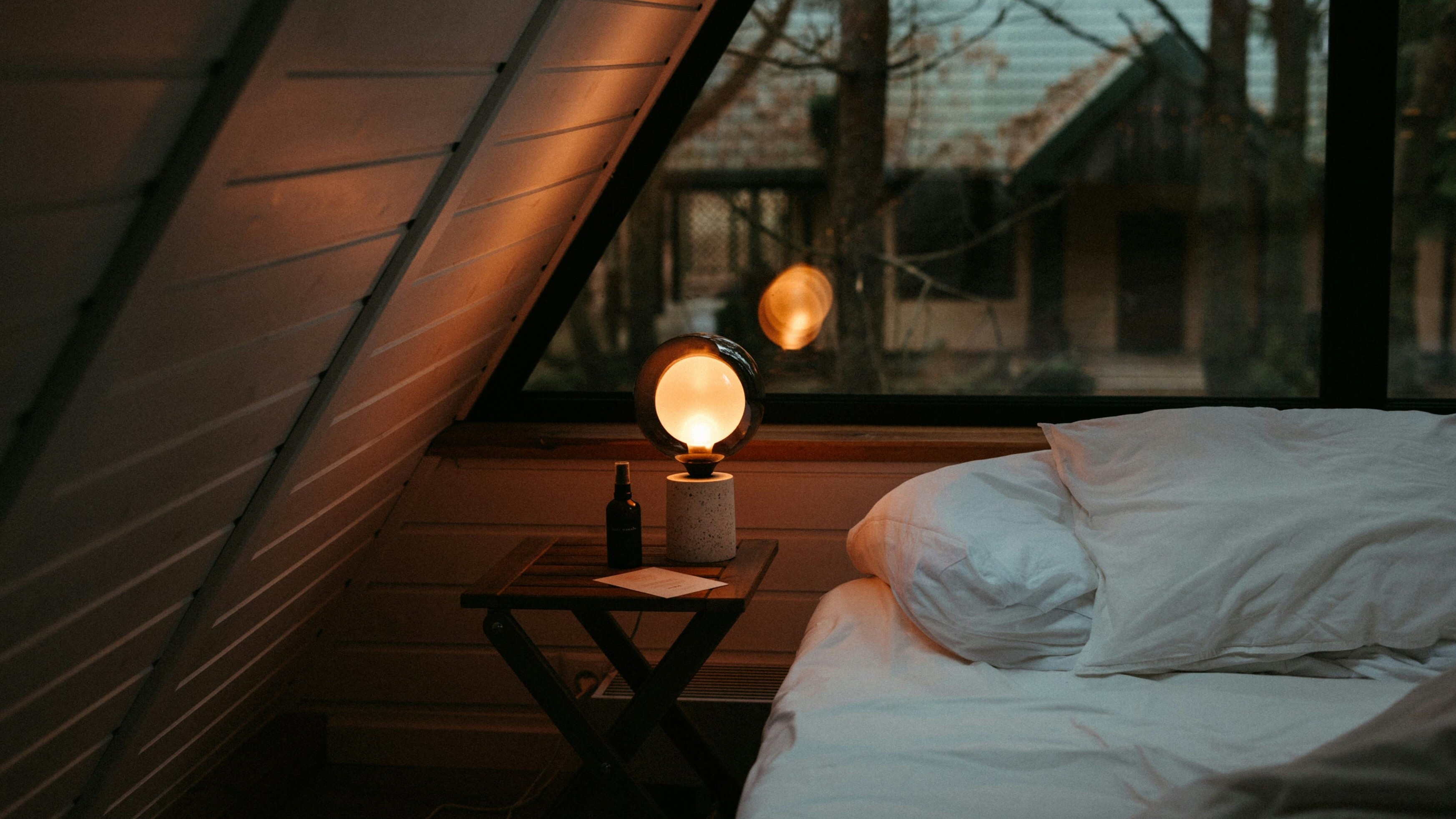 Ein gemütliches Dachzimmer mit weißer Bettwäsche und einer warm leuchtenden Lampe auf einem Holznachttisch schafft eine ruhige, angenehme Atmosphäre.