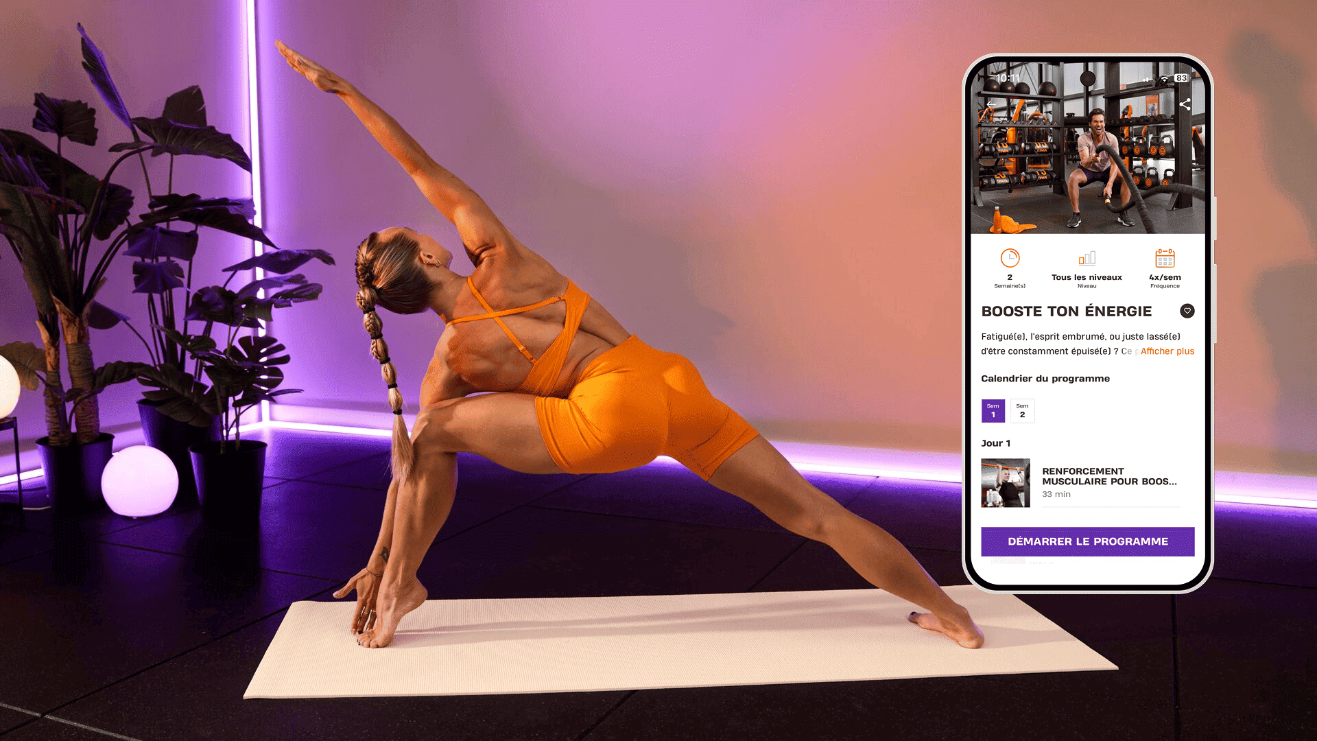 Une femme effectue une posture de yoga sur un tapis dans une pièce éclairée par des lumières violettes et entourée de plantes tropicales. Elle porte une tenue de sport orange vif. À côté d’elle, un smartphone affiche le programme Basic-Fit “Booste ton énergie”, proposant des séances pour renforcer la force et la confiance en soi.