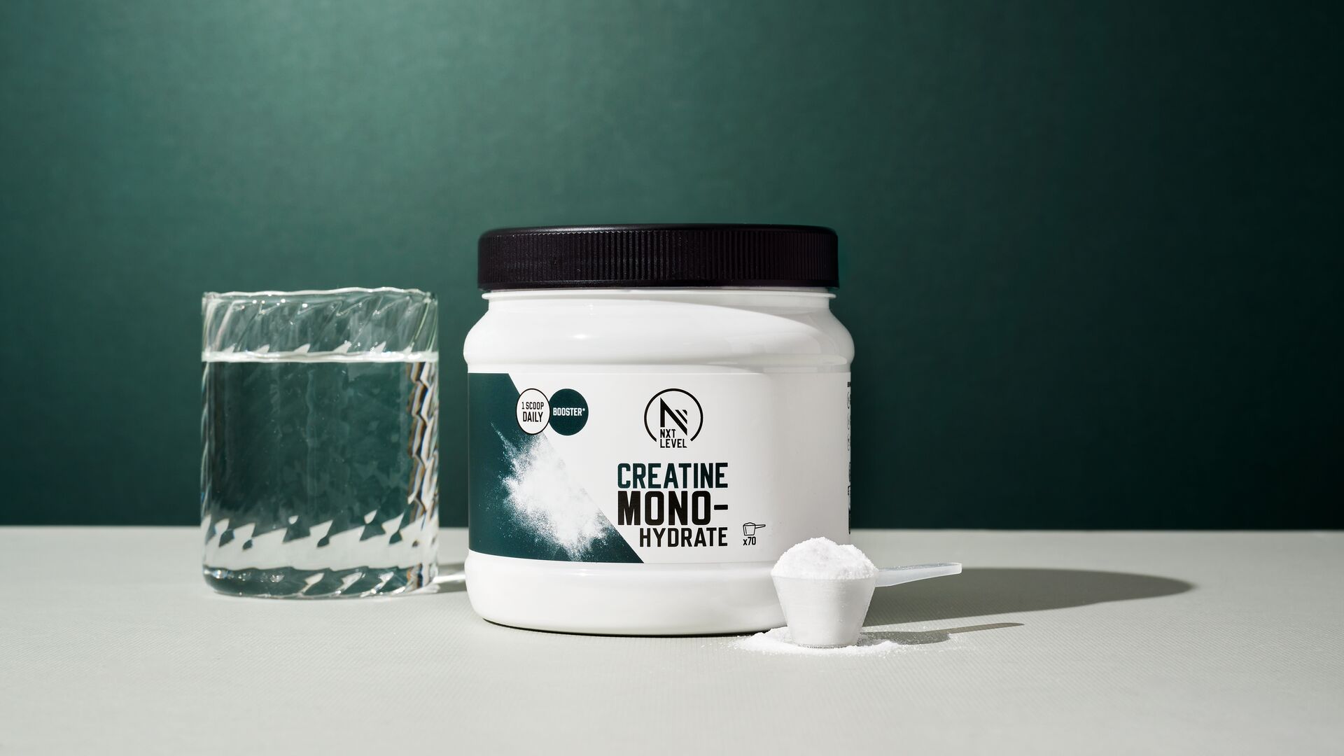 Een pot creatine monohydraat staat op een tafel naast een glas water en een maatschep met poeder, gefotografeerd tegen een donkergroene achtergrond.
