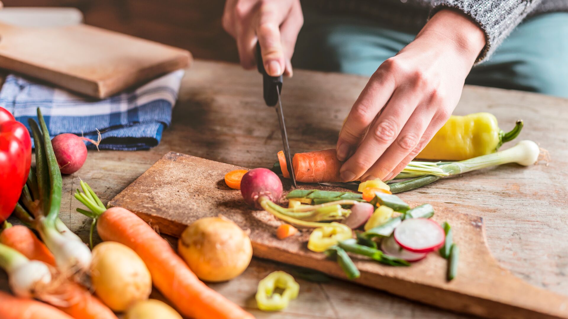Close-up van handen die verse groenten snijden op een houten snijplank, omringd door kleurrijke ingrediënten zoals wortels, aardappels, radijsjes en paprika's.