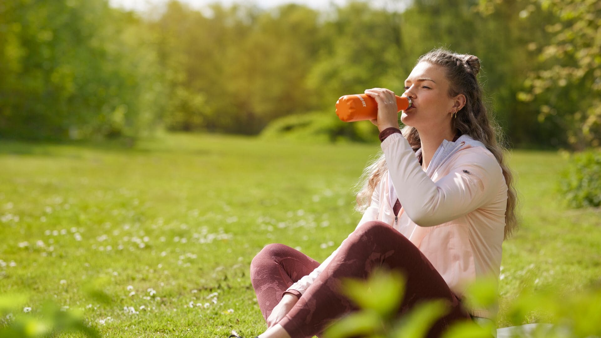 Eine Frau in einer leichten Jacke und bordeauxroter Leggings sitzt im Gras in einem sonnigen Park und trinkt aus einer orangefarbenen Trinkflasche nach ihrem Workout.
