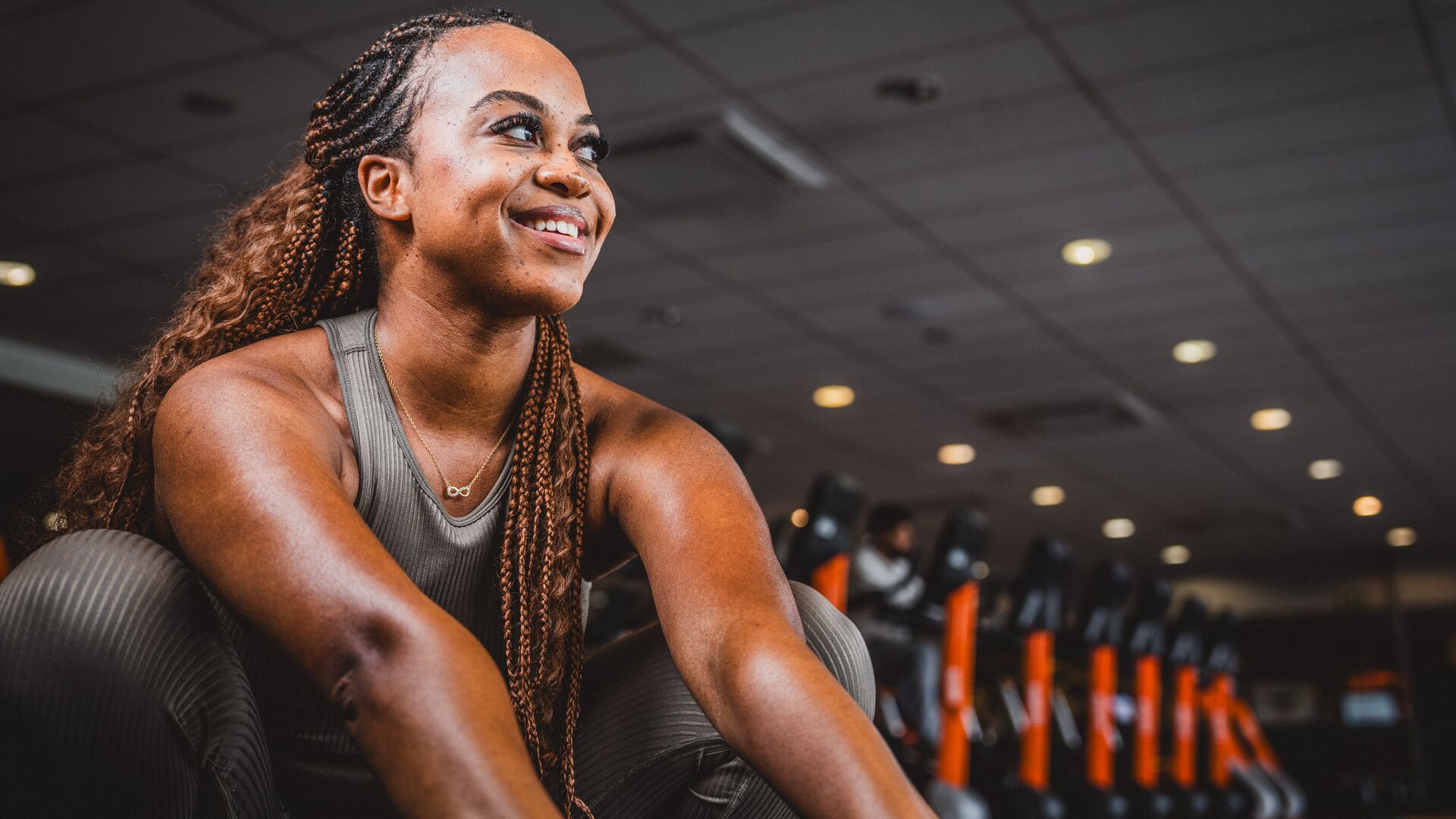 Woman in a gray sports outfit sitting on the gym floor, smiling, with cardio machines and orange accents in the background.