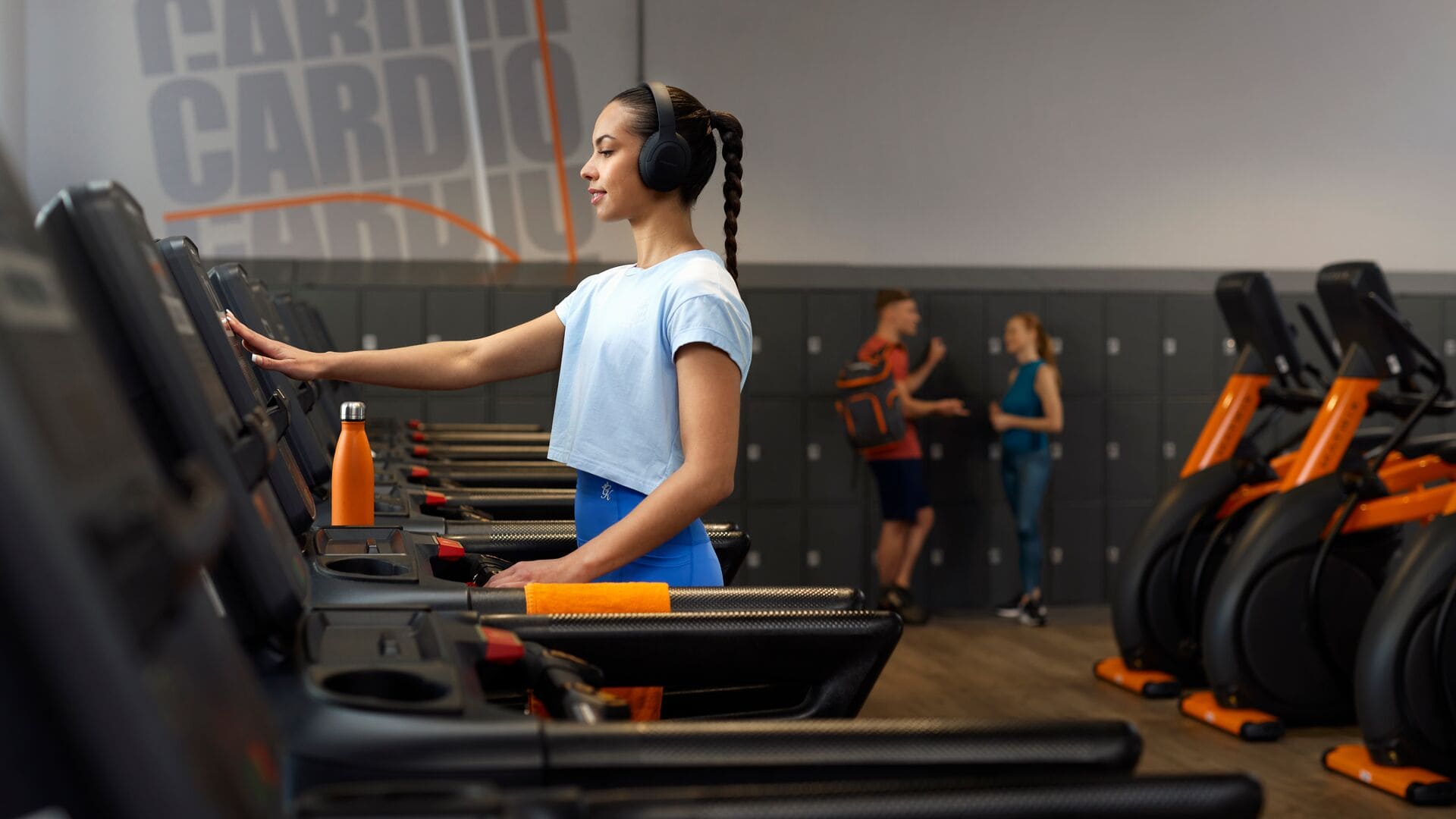 Eine Frau in einem hellblauen Top und einer blauen Leggings trainiert auf einem Laufband bei Basic-Fit, trägt Kopfhörer, mit einer orangefarbenen Trinkflasche davor.