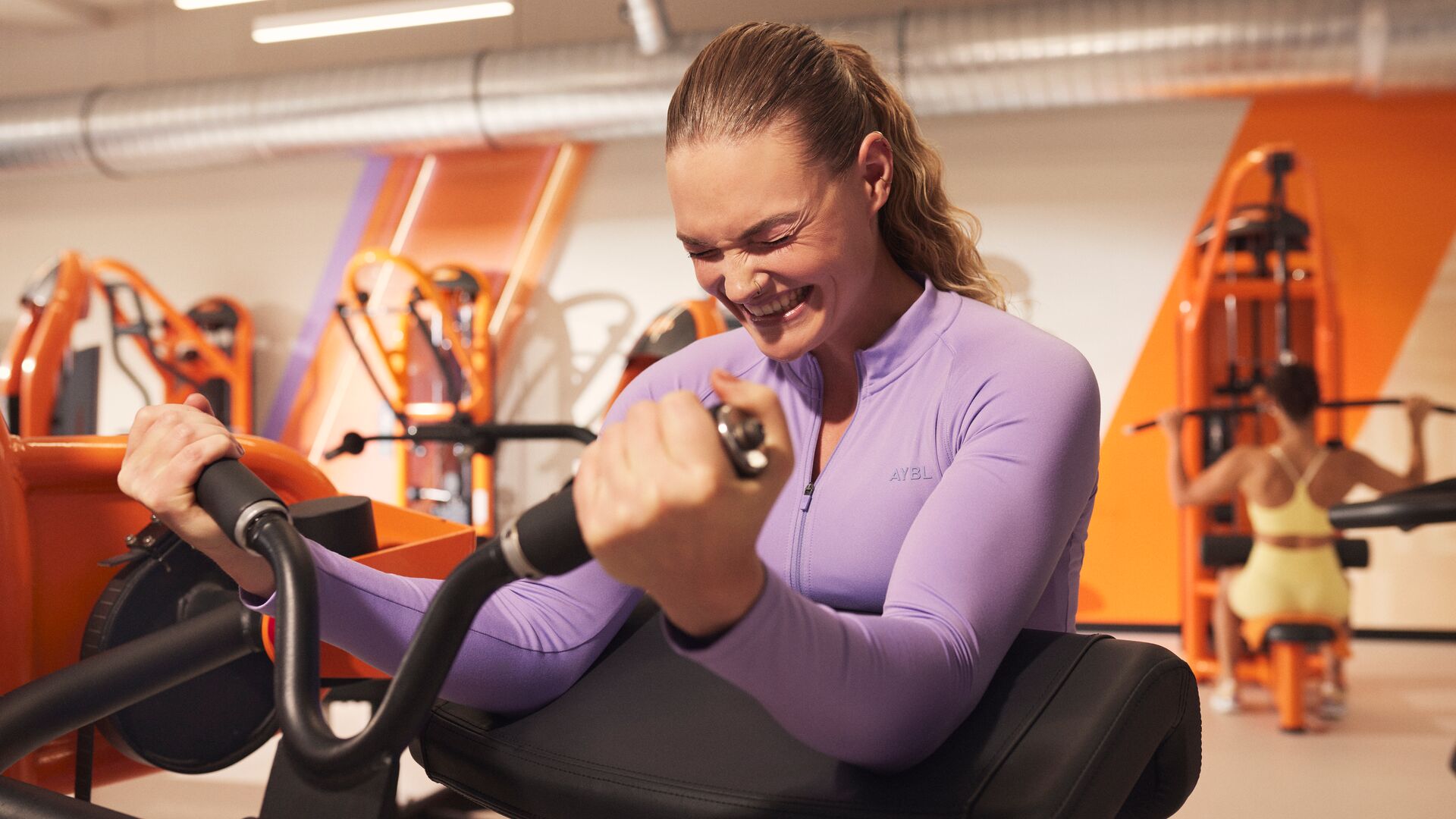 Een vrouw voert een zittende krachtoefening uit op een toestel in een Basic-Fit club en glimlacht terwijl ze zich op haar training concentreert. Krachttoestellen en oranje accenten zijn zichtbaar op de achtergrond.
