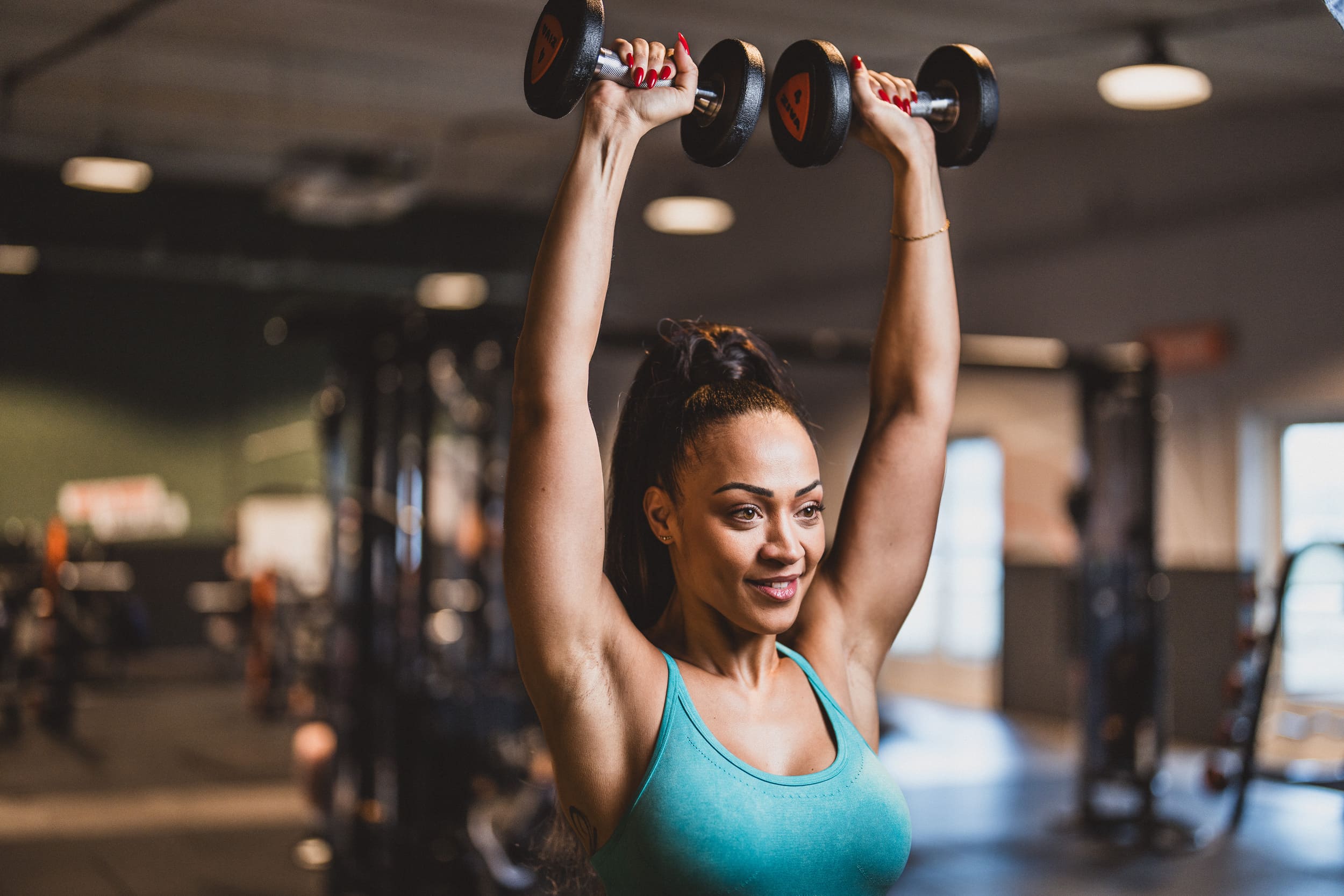 Eine Frau in einem türkisfarbenen Sporttop hebt selbstbewusst Kurzhanteln über den Kopf in einem hell erleuchteten Basic-Fit-Studio, umgeben von Trainingsgeräten und einer energiegeladenen Atmosphäre.