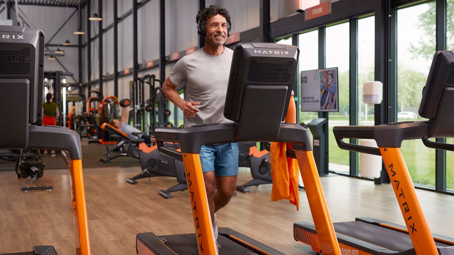 Ein Mann mit grauem T-Shirt und blauer Shorts läuft auf einem Laufband in einem Basic-Fit-Fitnessstudio. Er trägt Kopfhörer und lächelt, während andere Mitglieder im Hintergrund trainieren.