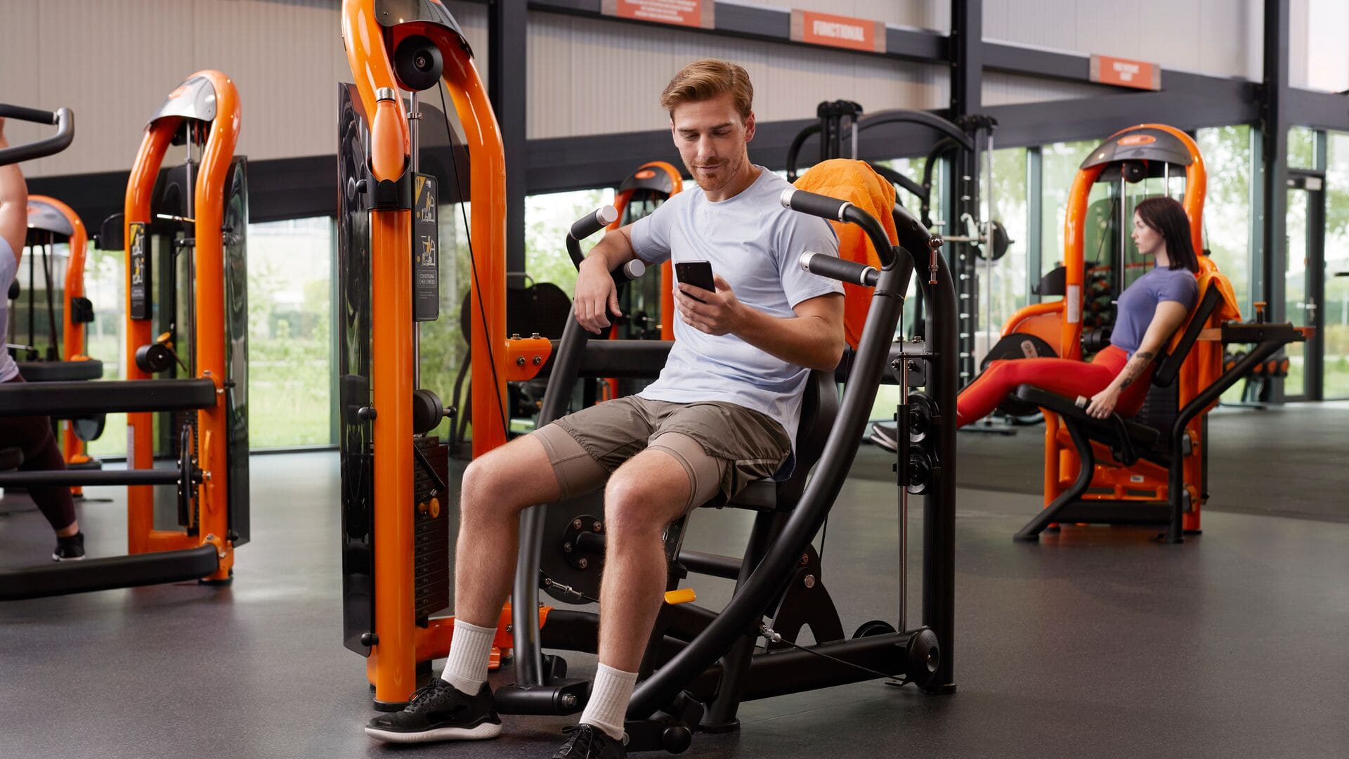 Mann sitzt an einer Brustpresse-Maschine und schaut auf sein Handy im Basic-Fit Fitnessstudio, während andere Mitglieder im Hintergrund Kraftgeräte benutzen.
