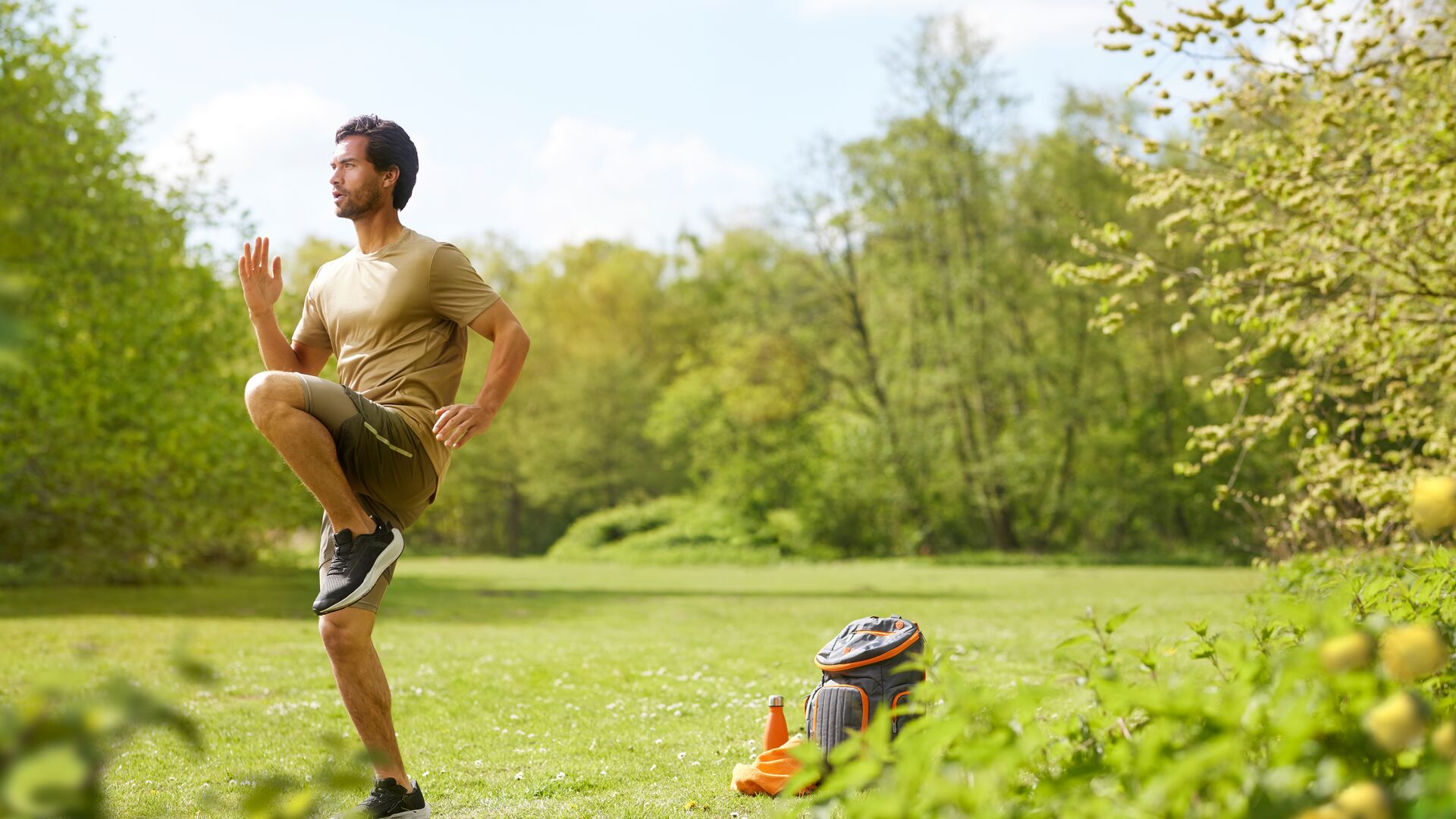 Man doet een hoge-knieën cardio-oefening in een zonnig park, met een rugzak, drinkfles en handdoek in het gras.