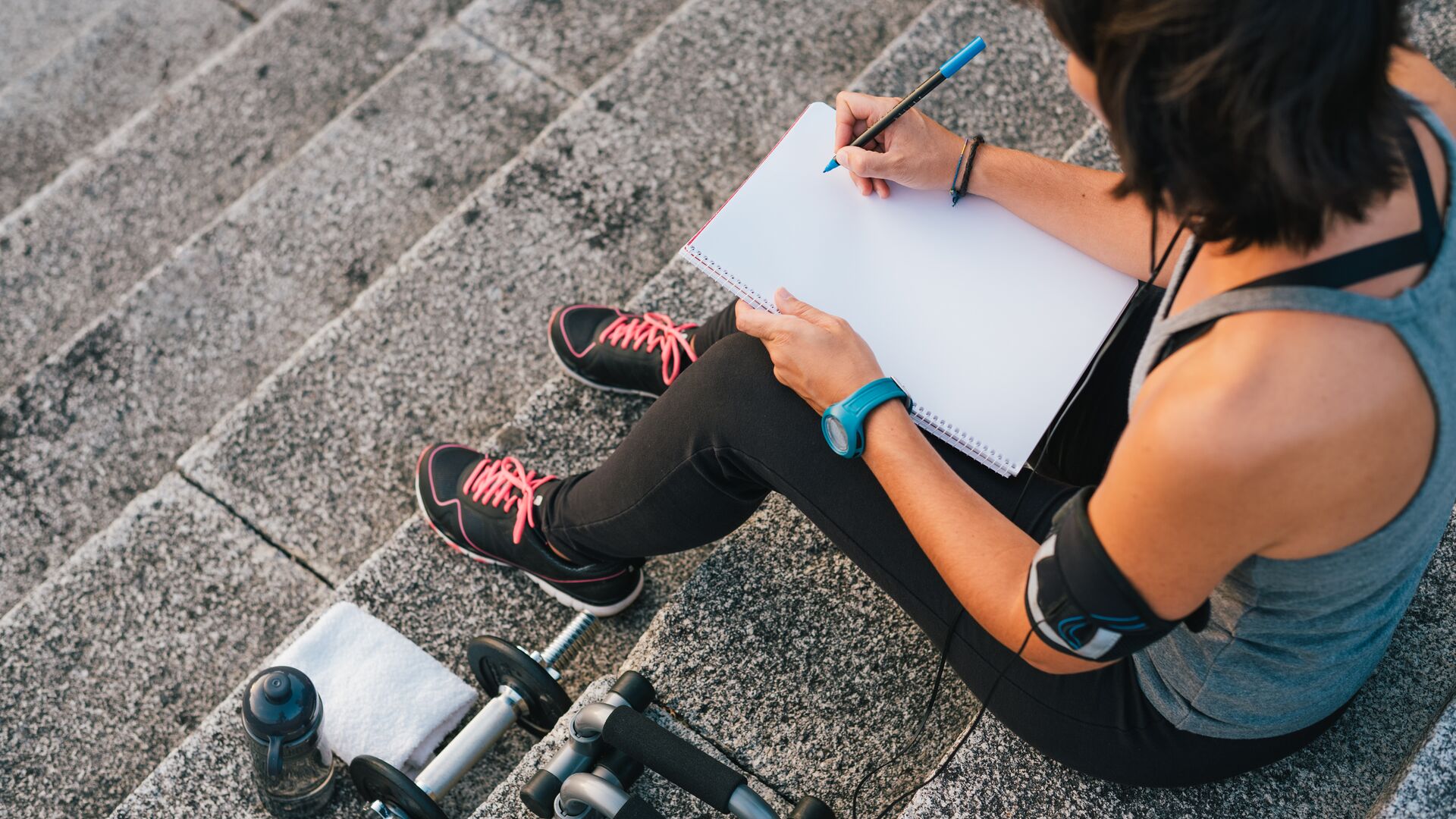 Woman in workout gear sitting on outdoor concrete steps with a notebook on her lap, ready to set her workout goals. Next to her: a towel, dumbbells and a water bottle — ready to go.