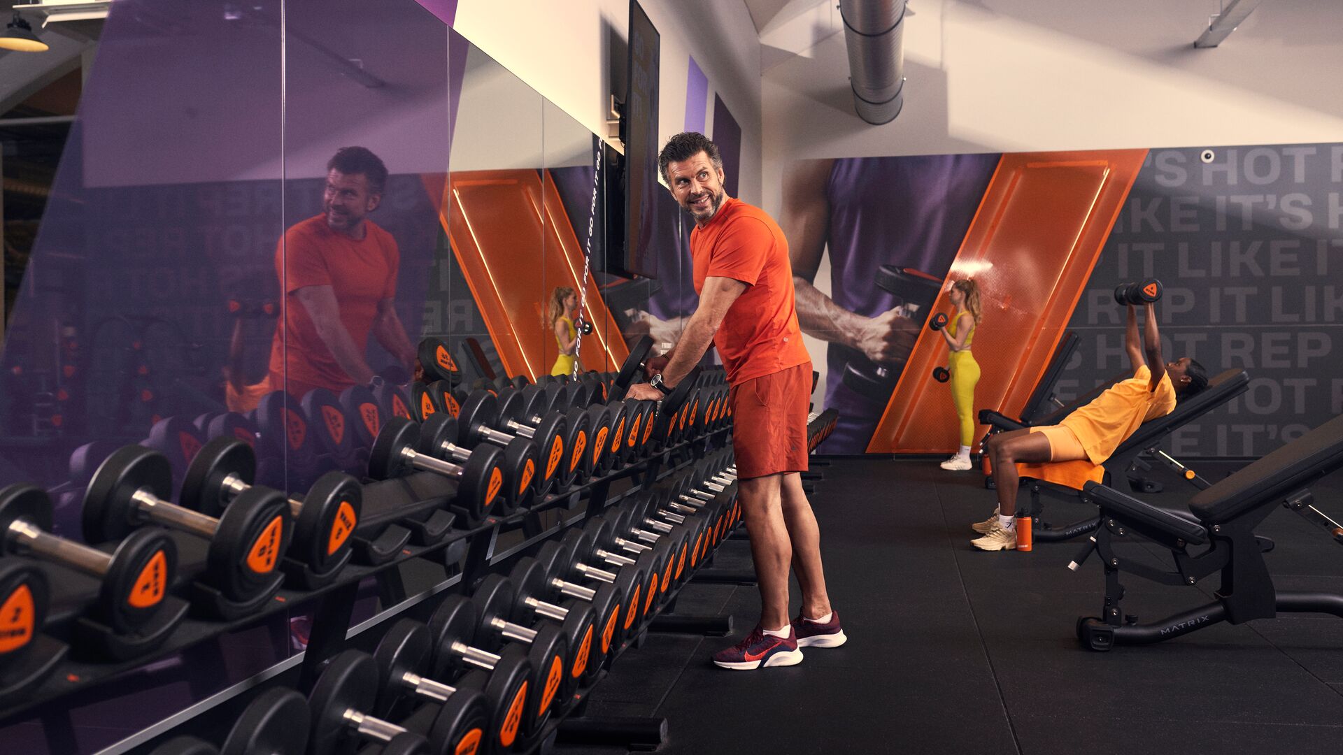 Mann wählt Kurzhanteln im Fitnessstudio, während im Hintergrund ein anderes Mitglied auf einer Bank trainiert