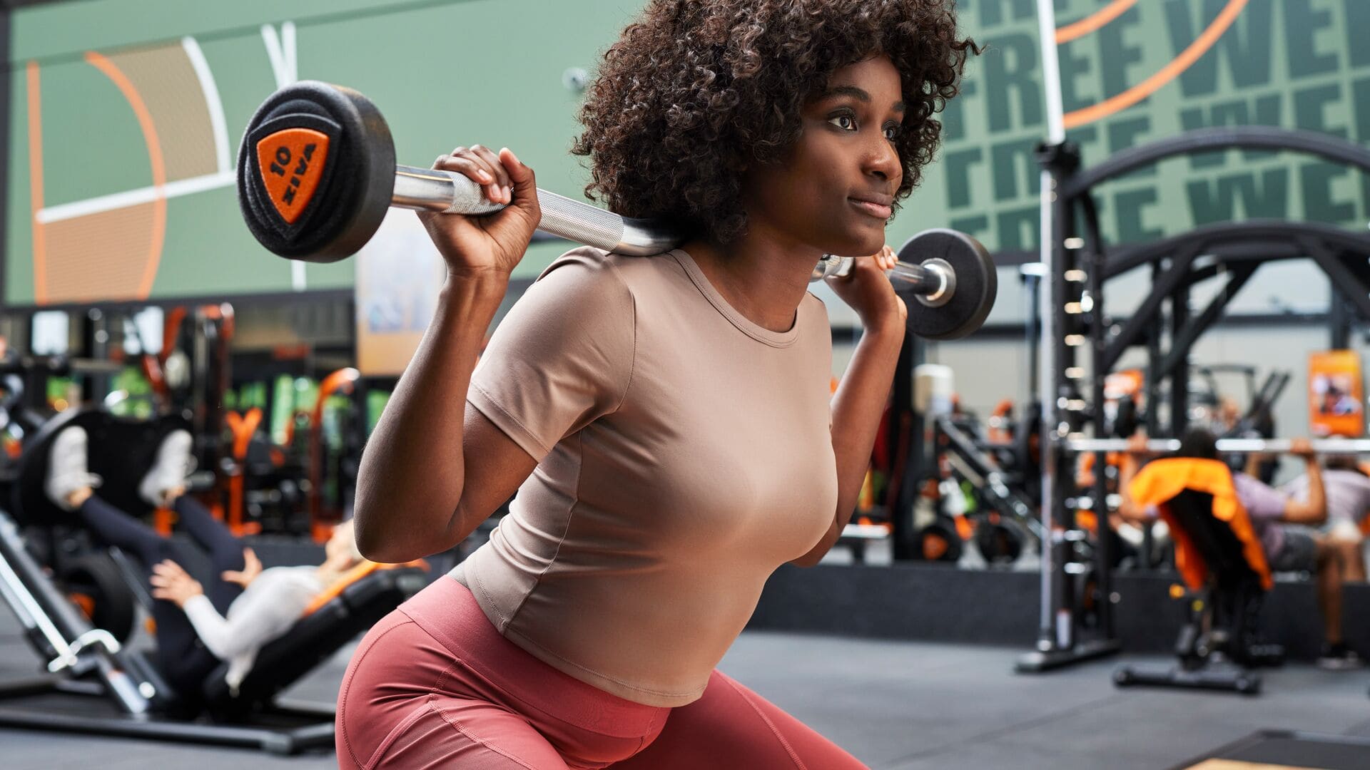 Eine Frau macht eine Kniebeuge mit einer Langhantel auf den Schultern in einem Basic-Fit-Studio, fokussiert und selbstsicher in einer dynamischen Trainingsumgebung mit anderen Mitgliedern im Hintergrund.