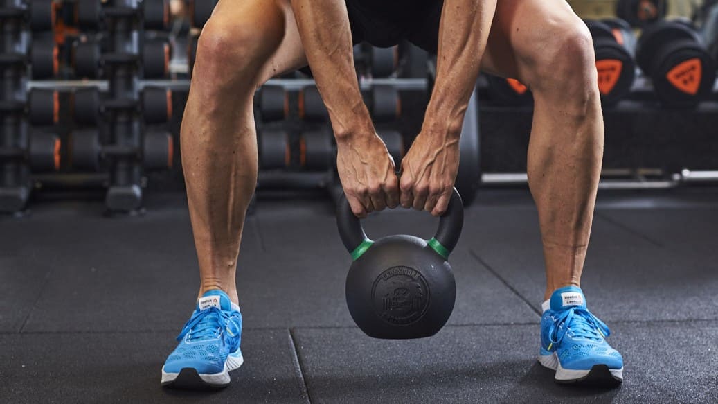 Personne soulevant une kettlebell dans une salle Basic-Fit, portant des chaussures de sport bleues, entourée d’haltères.